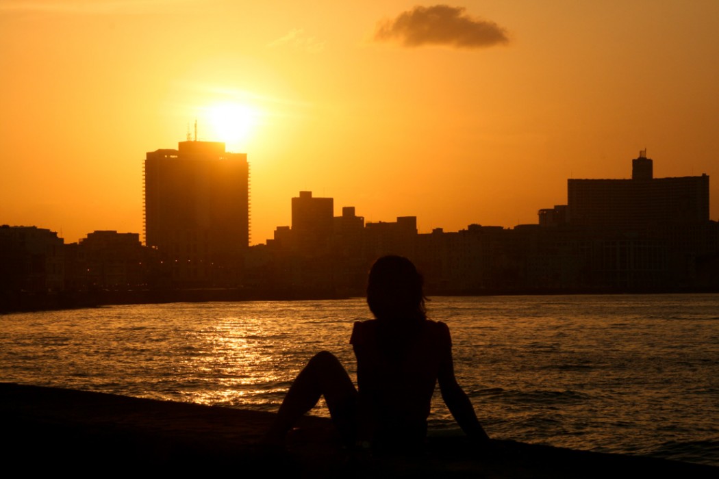 A young woman sits on the Malecón wall as the sun sets over the bay, creating a silhouette of her and the buildings