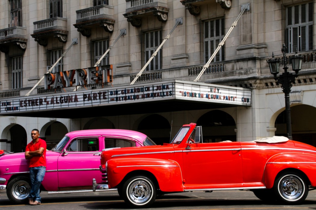 A man stands next to his classic pink car, itself sitting parked next to an old red convertible.