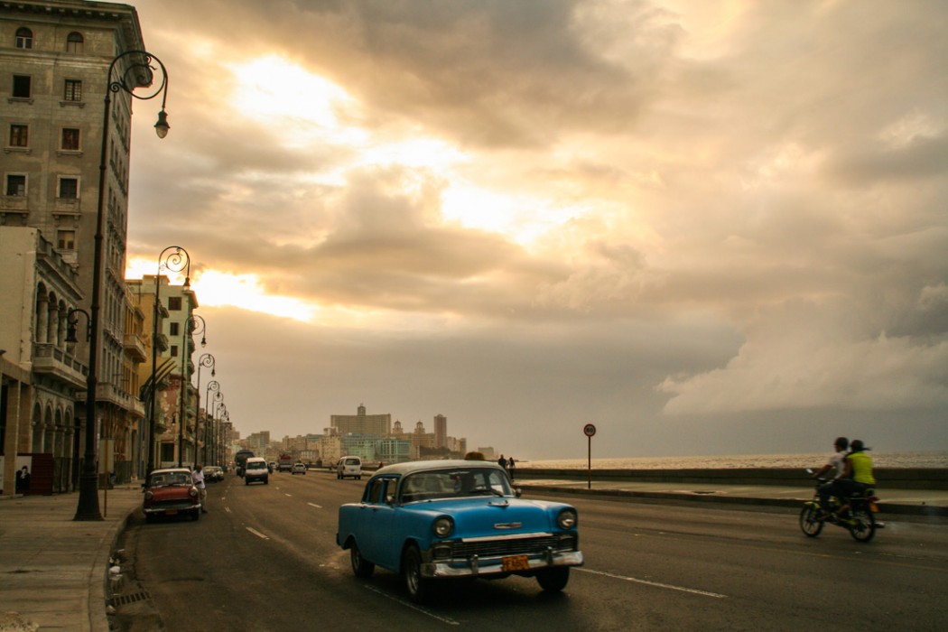 Classic cars along the Malecón A blue chevrolet with the eagle ornament flies down the Malecón as the sun lights up the clouds behind