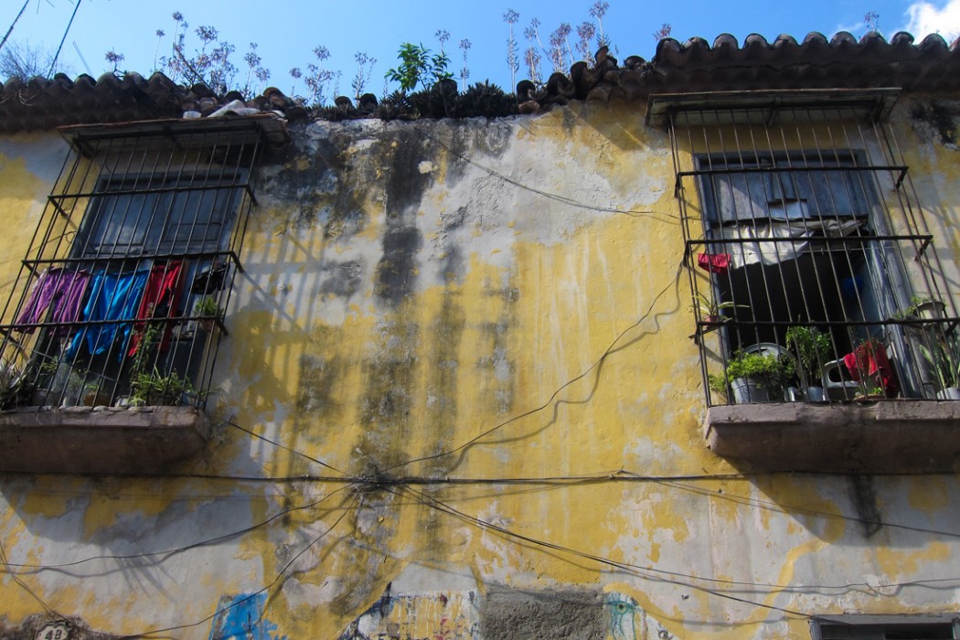 A picture of the second story of a faded yellow house, with a part of the roof missing and weeds growing on the remainder