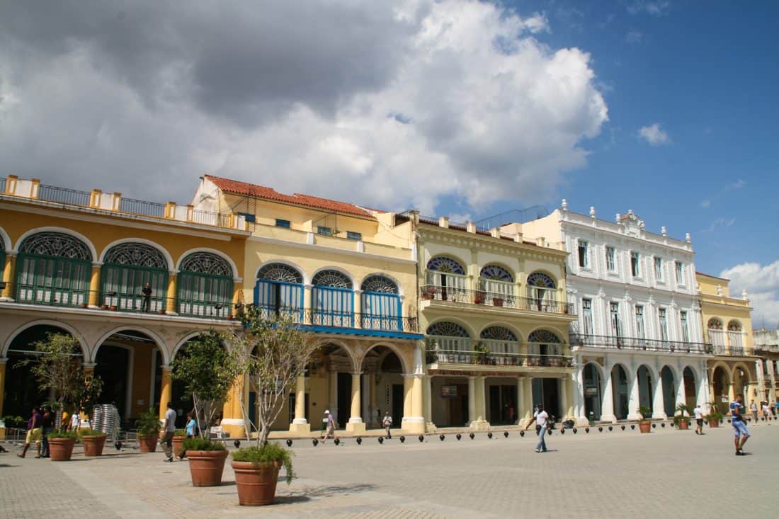 A shot across a tree-lined plaza with beautifully painted buildings on the far edge