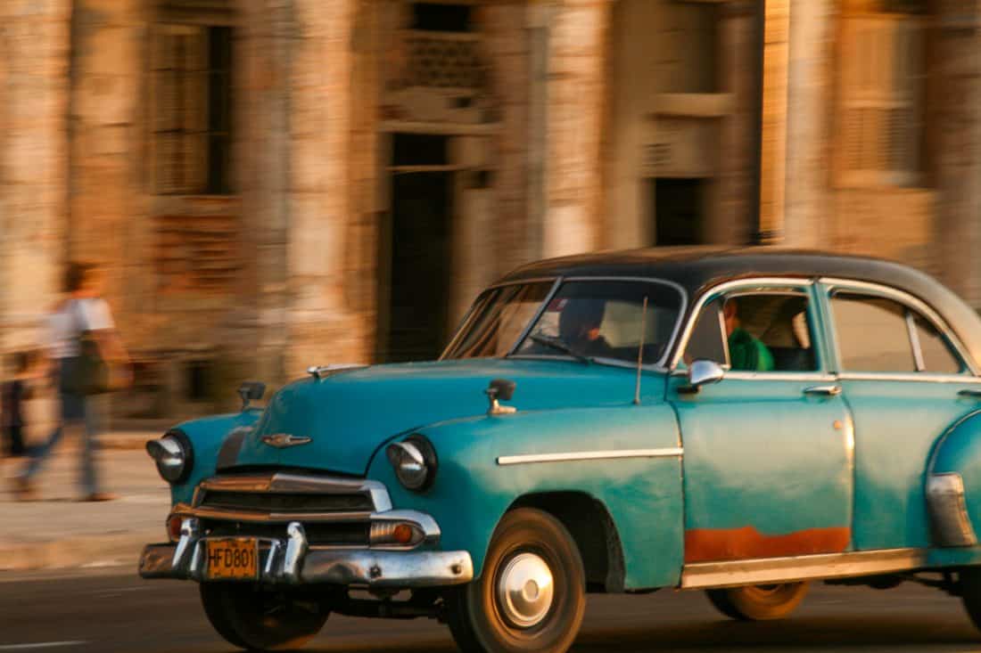 A turquoise car races along the Malecón with the sun turning the old building behind it orange
