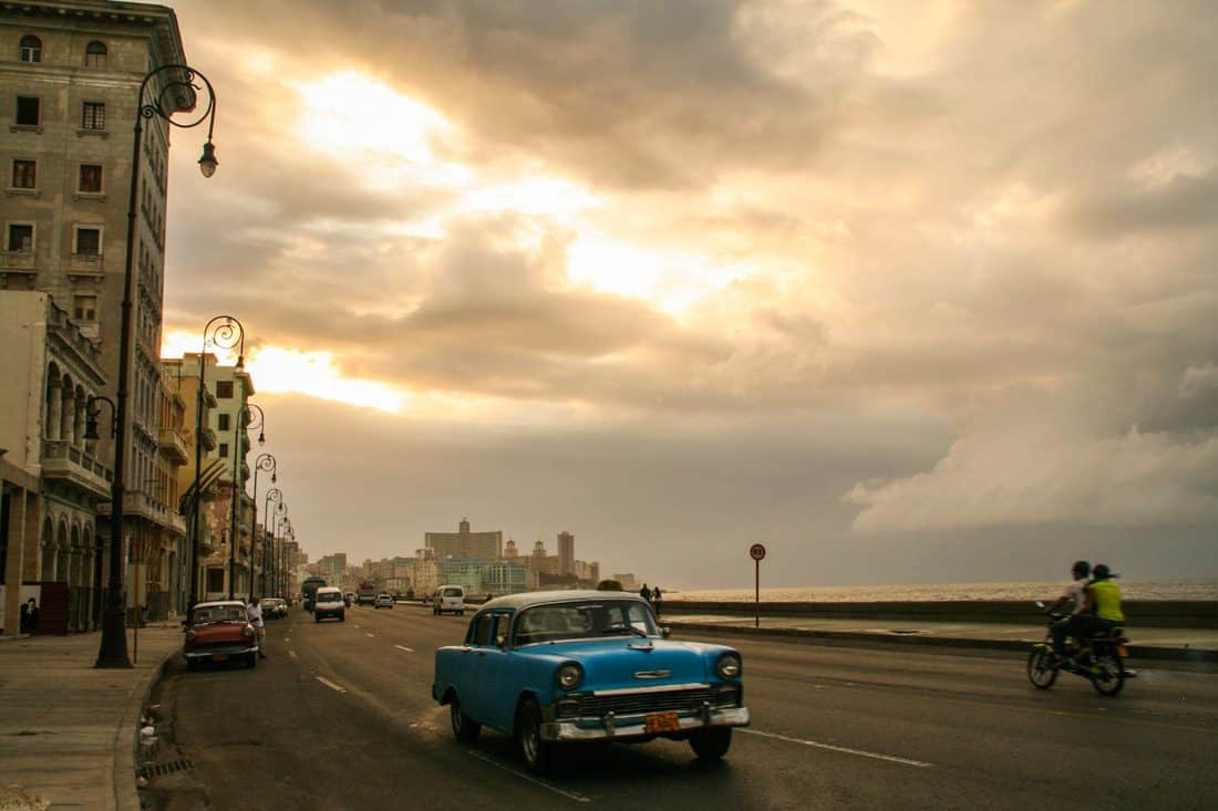 A blue chevrolet with the eagle ornament flies down the Malecón as the sun lights up the clouds behind
