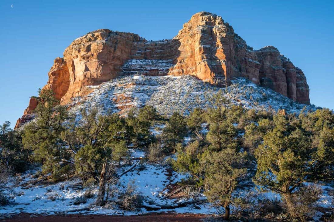 Large red rock formation of Courthouse Butte in Sedona sprinkled with snow with green trees below