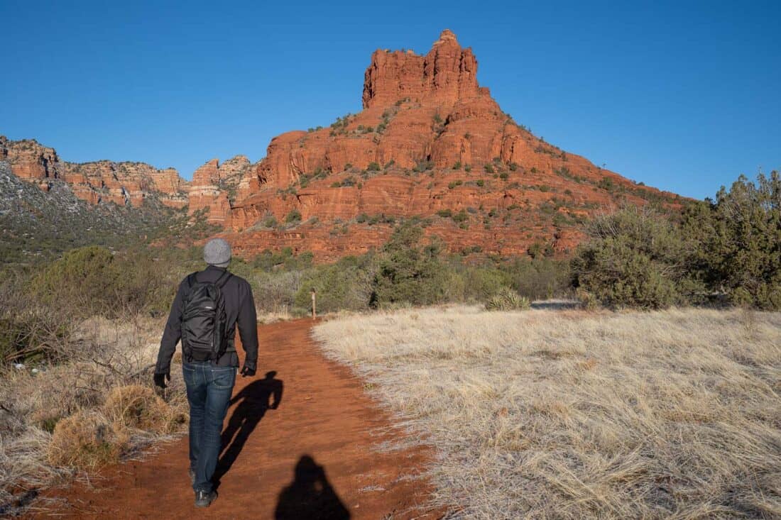 Simon walking towards distinctive red Bell Rock in Sedona along a red sand path surrounded by dried grass and green trees, Arizona, USA