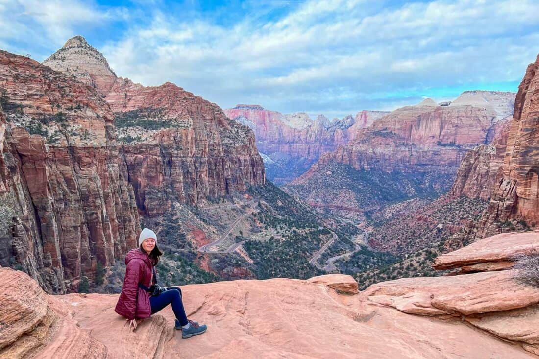 Erin perched at the edge of the Canyon Overlook with a panorama of pink cliffs, a winding hiking trail, and cloudy blue sky, Zion National Park, Utah, USA