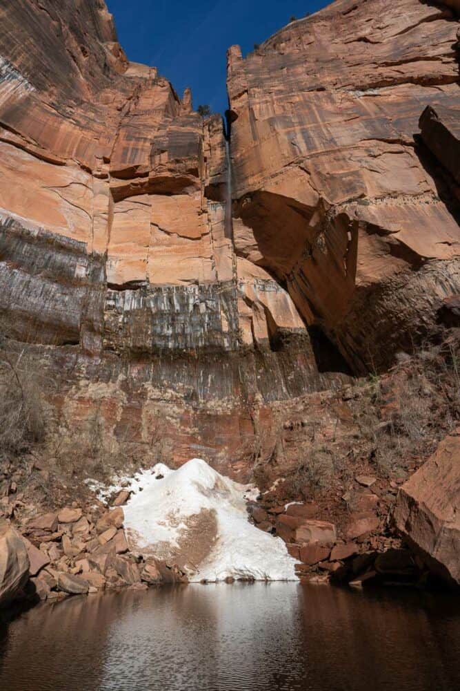 Upper Emerald Pool in Zion National Park, with towering pink cliffs and thin waterfall cascading down to a small snow drift