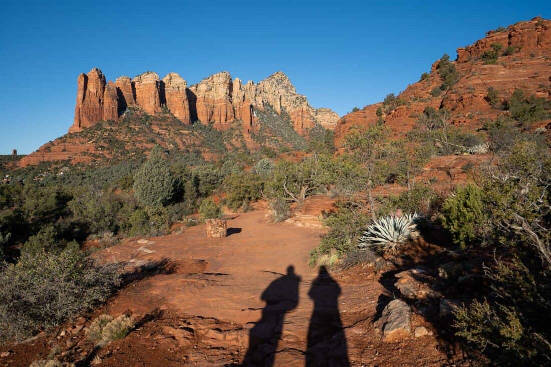 Early morning shadows on smooth red path on the Jordan Trail in Sedona, craggy rocks in the distance with blue sky and greenery