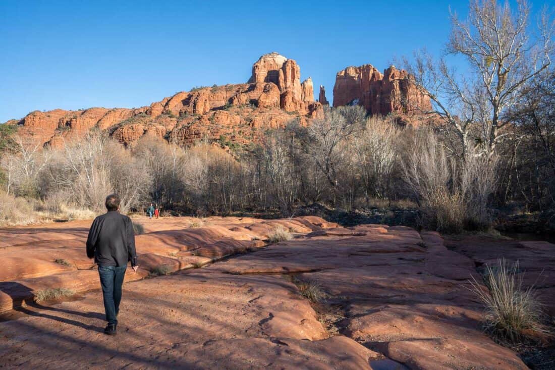 Simon walking along red flat rock towards Cathedral Rock looming above trees, Sedona, Arizona, USA