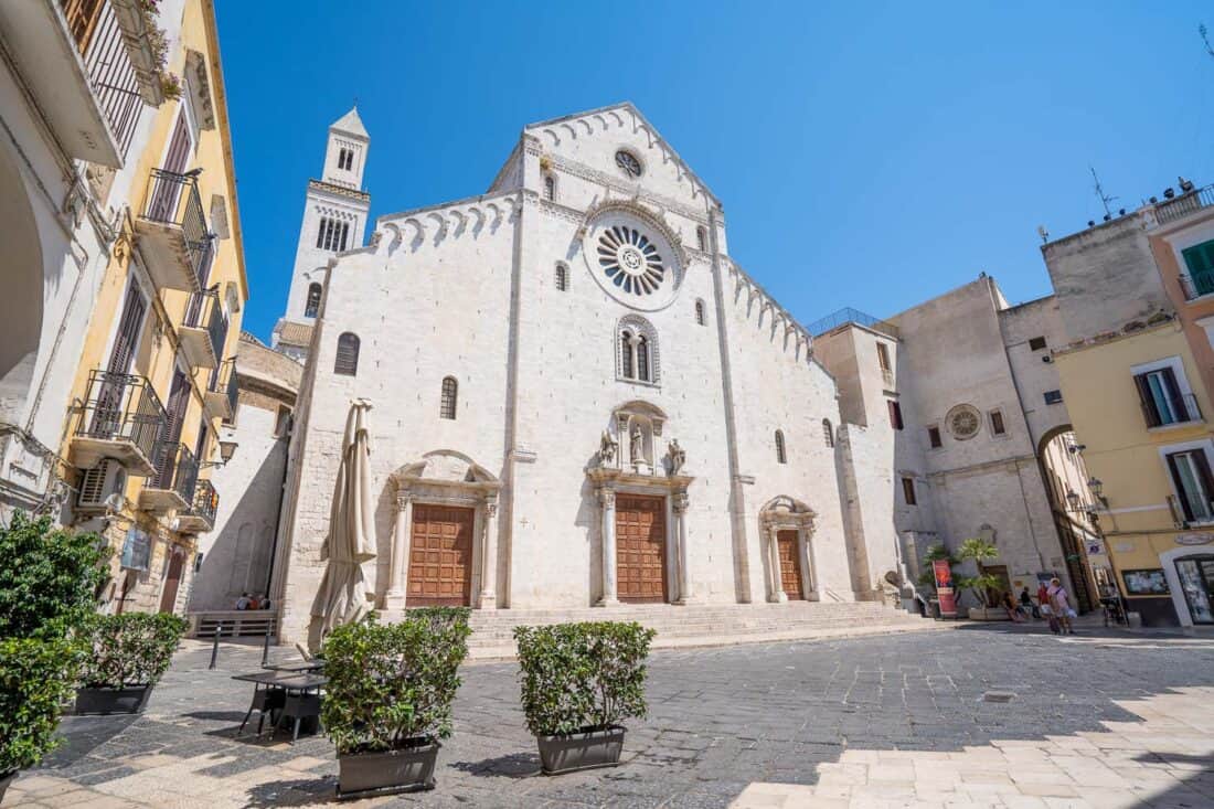 White facade of Il Duomo overlooking a small piazza with potted plants and yellow painted buildings