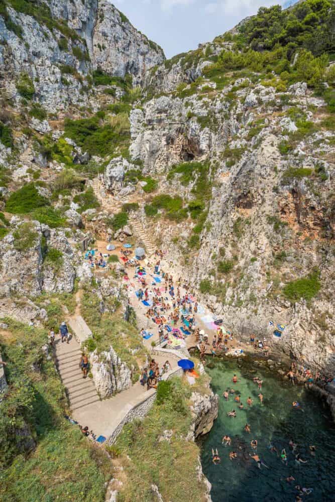 View from above of the busy scenic cove of Il Ciolo with stone steps leading down to the cliffs, Salento