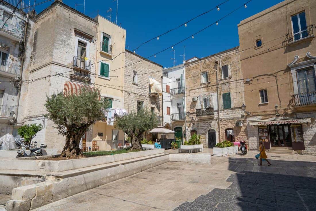 Woman walking across a quiet Largo Albicocca in Bari with small trees, restaurants, and unlit lights strung above