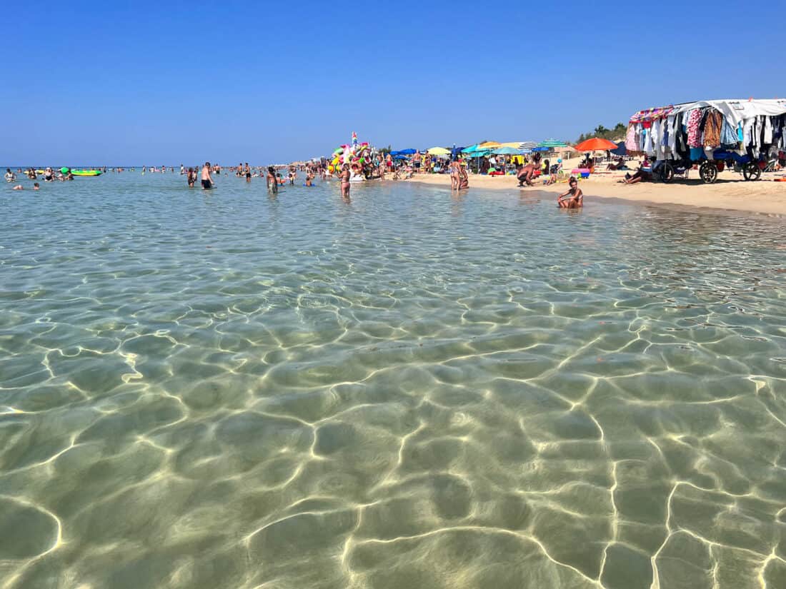Busy beach at Marina di Pescoluse with people in the shallow clear water and vendors along the sand