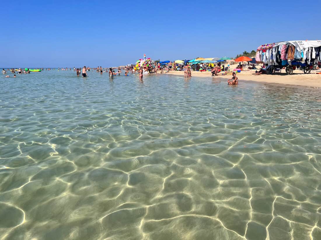 Busy beach at Marina di Pescoluse with people in the shallow clear water and vendors along the sand, Puglia, Italy