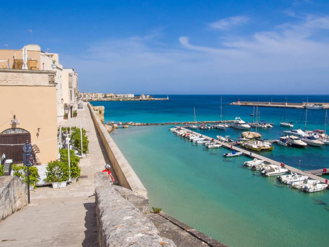 Path leading down to Salento harbour with boats moored in clear blue waters on a sunny day