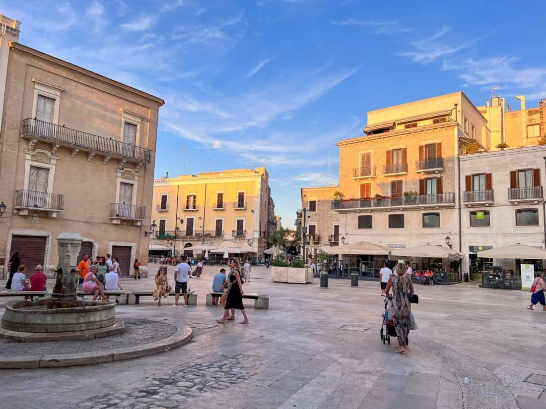 People walking and sitting on stone benches in Piazza Mercantile, surrounded by old buildings and bars 
