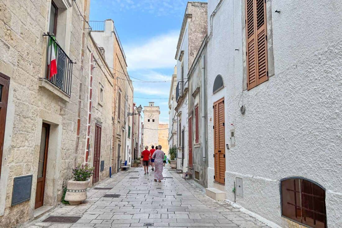 People walking down paved narrow Via Roma, lined with pale stone buildings with wooden shutters, Castro, Italy 