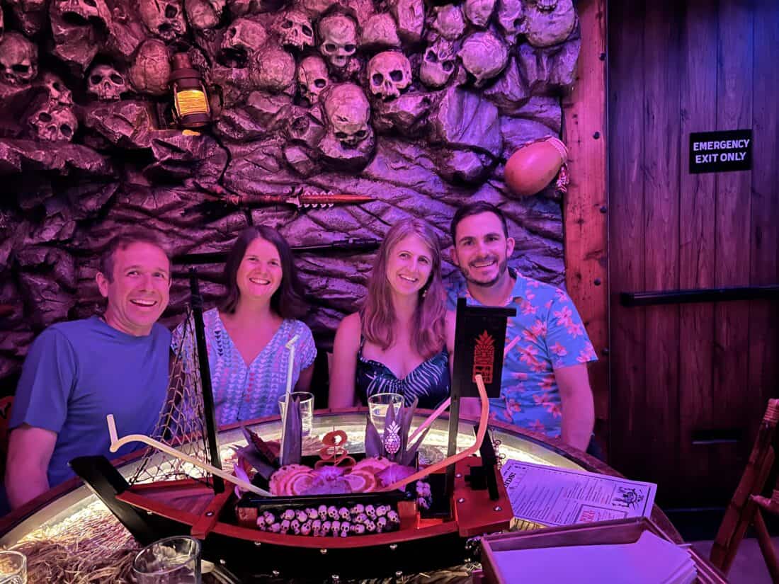Simon, Erin, Tom, and Jenny sitting in front of a wall of skulls with a cocktail in front of them in the shape of a pirate ship