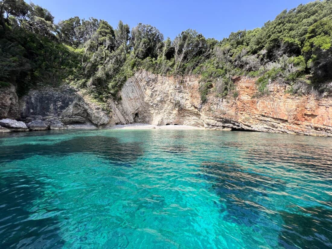 A photo from a boat looking back towards a white sand beach with large tree-covered cliffs behind it. The water is an iradescent blue.