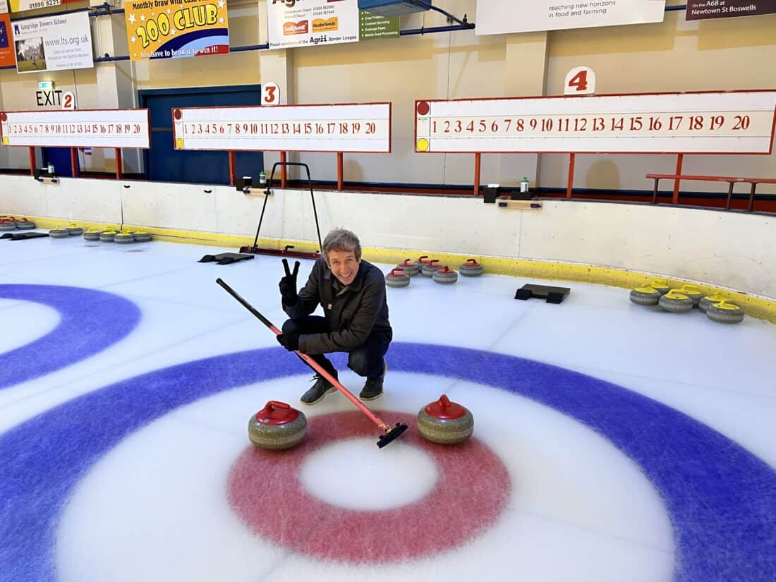 Simon on the target of a curling rink with two stones very close to the centre. He is kneeling down and giving a V sign.