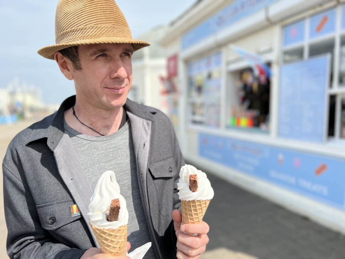 A portrait of Simon in front of Worthing Pier holding two 99s