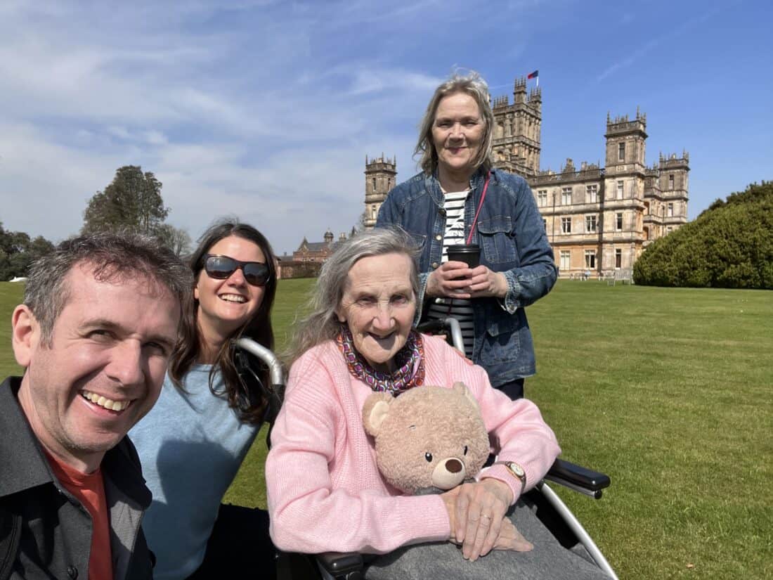 Simon, Erin, Erin's Nan, and Erin's Aunt standing in front of Highclere Castle. Erin's nan is in a wheelchair holding a teddy bear.