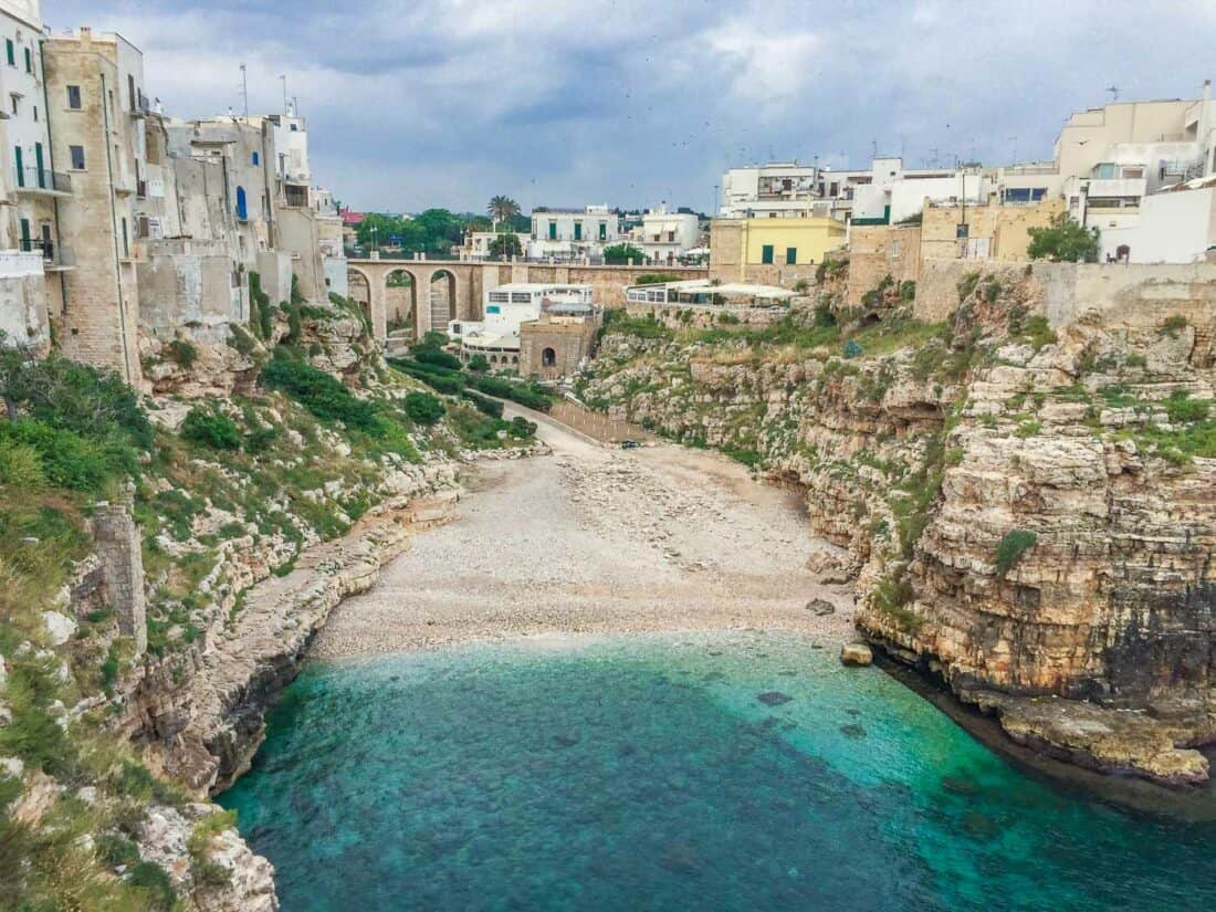 Quiet Lama Monachile beach in May surrounded by buildings perched on craggy cliffs and backed by a tall viaduct, Puglia