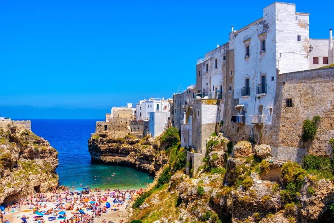 Crowded Polignano a Mare Beach in summertime, overlooked by buildings perched on a craggy cliff